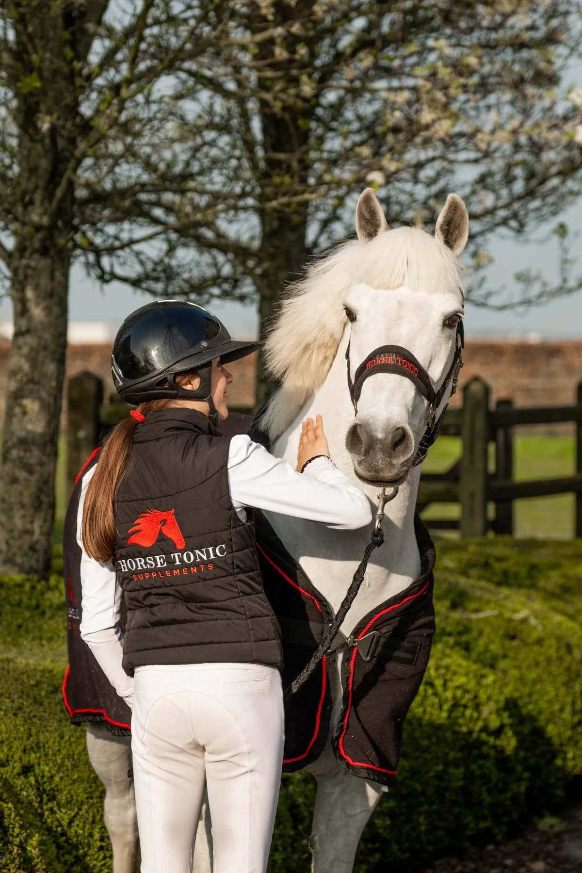 Ruiter in een Horse Tonic bodywarmer naast een wit paard bij de stal